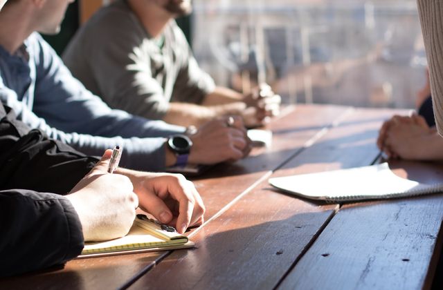 People around a wooden table in a meeting, one writing in a notebook