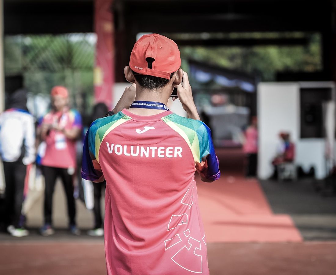 A volunteer in a cap and a pink 'Volunteer' shirt, seen from behind at an outdoor event